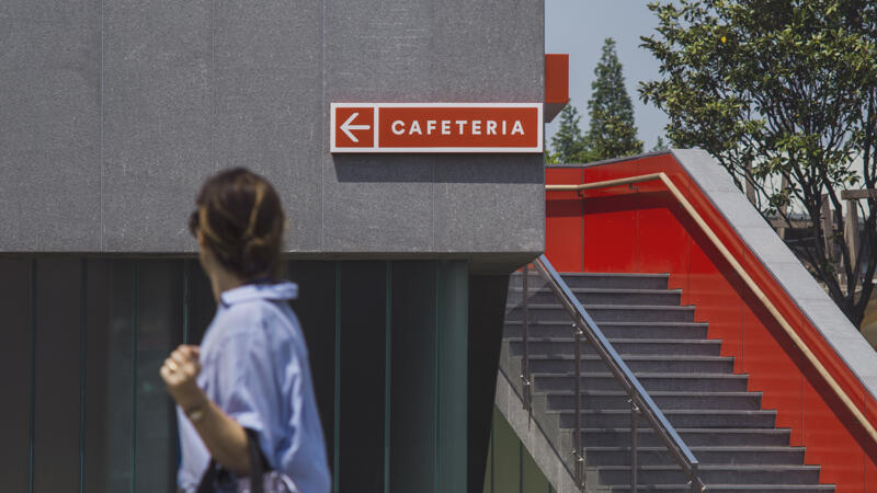 Wayfinding signage affixed to a concrete wall that reads "Cafeteria" with a person walking by